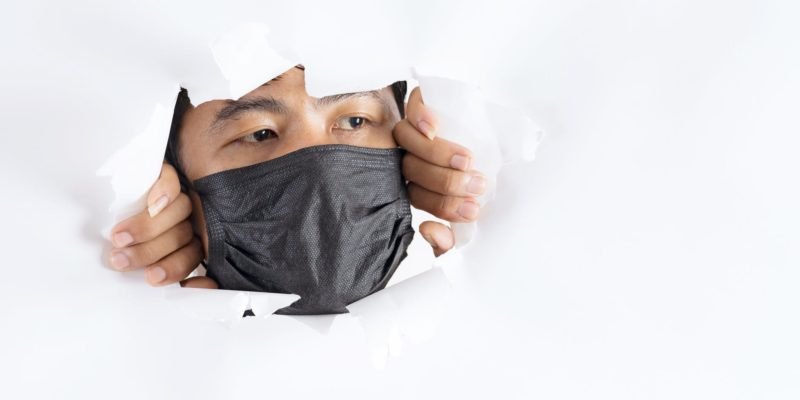 crop attentive man in face mask behind torn paper wall